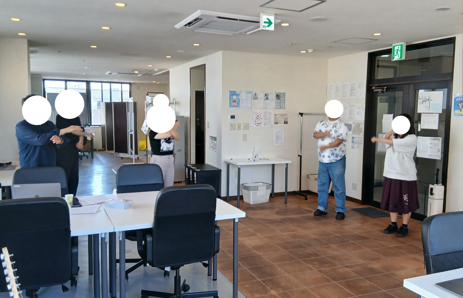 ALOHAの作業スペースで、机と椅子のそばに立ってラジオ体操をする人たち