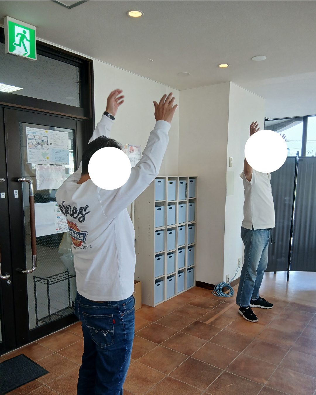 事業所の入口近くで、両腕を大きく上に伸ばしてラジオ体操をする2人
